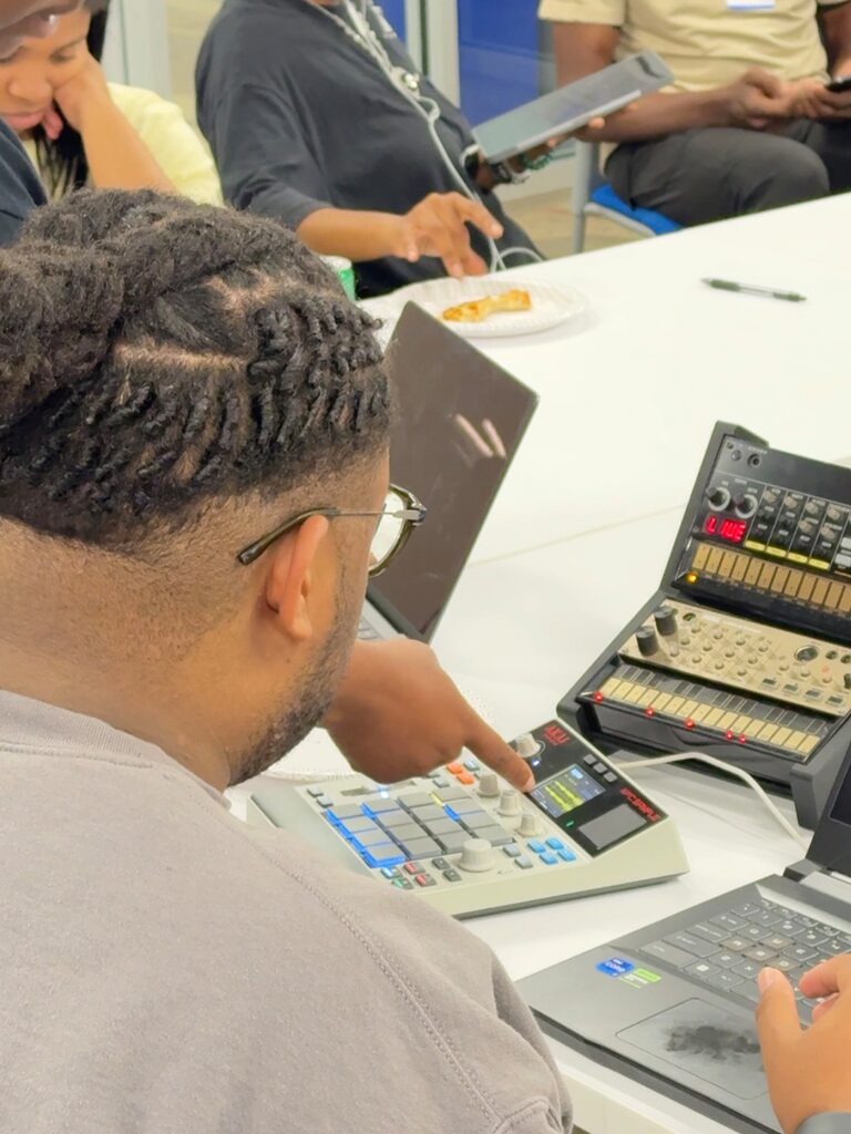 An attendee presses pads on a drum machine, next to a smaller synth and a laptop.
