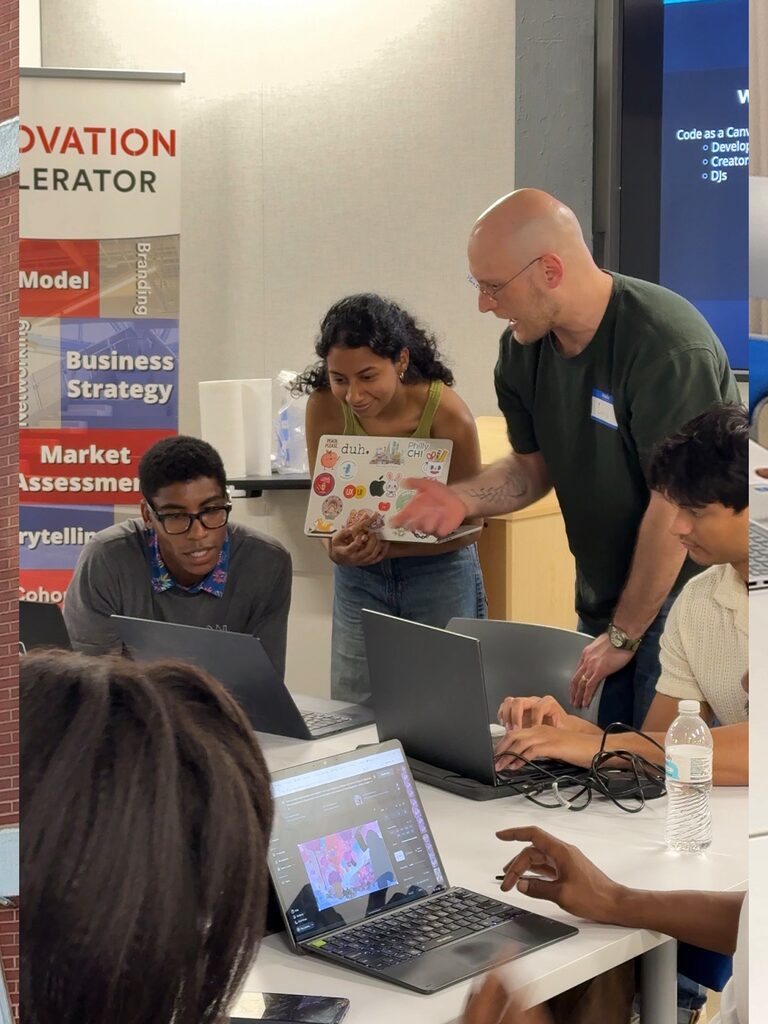 A helper leans over a table to point at a laptop while the people around it build.