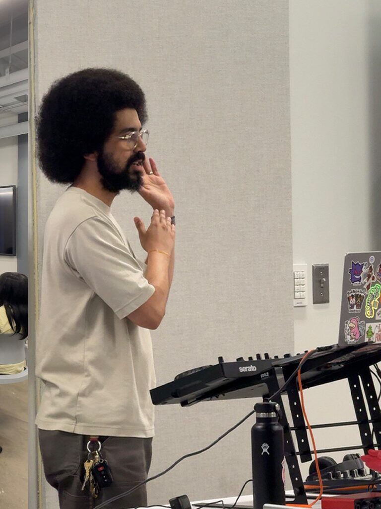 The presenter gestures while talking, beside a DJ controller on a stand.