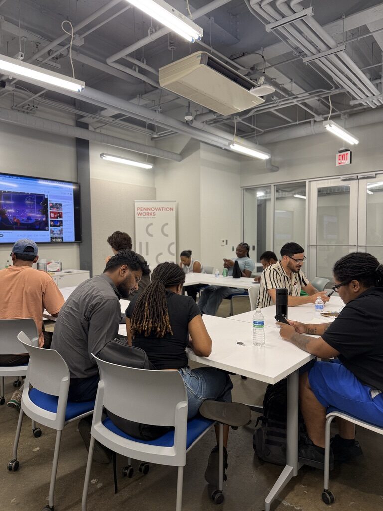 A wide view of the workshop mid-session: several tables of people building, presentation screens on the far wall.