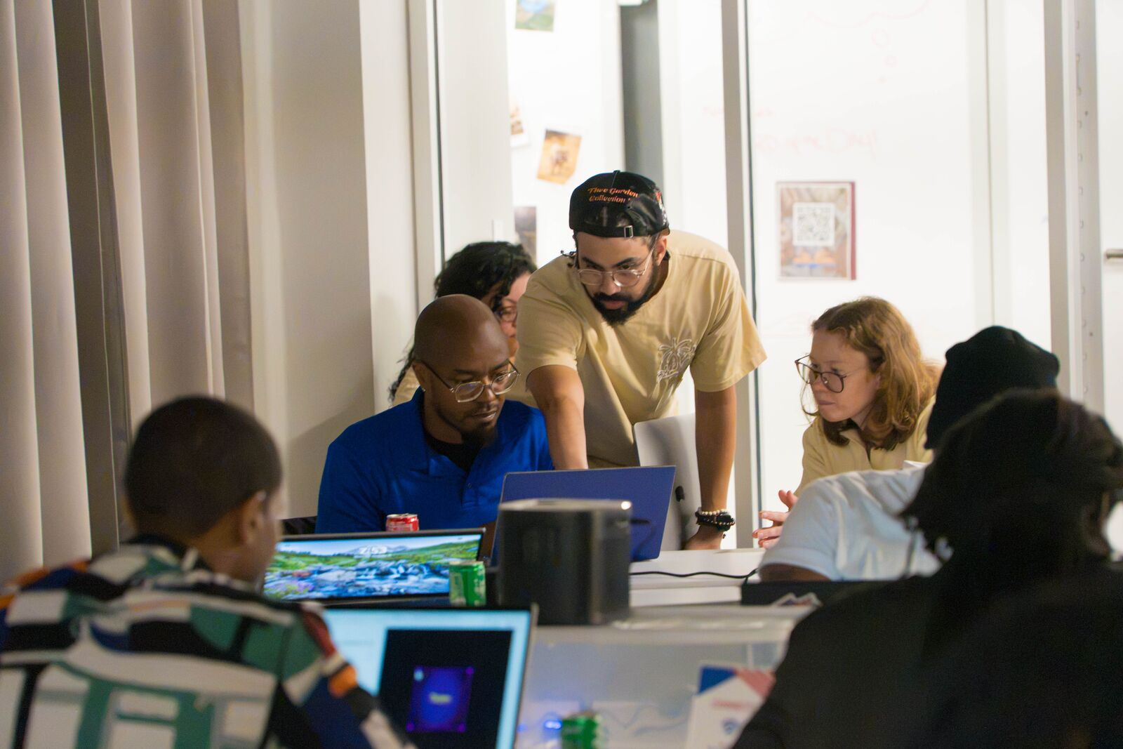 A facilitator in a cap leans over a table to help a group of attendees at their laptops.