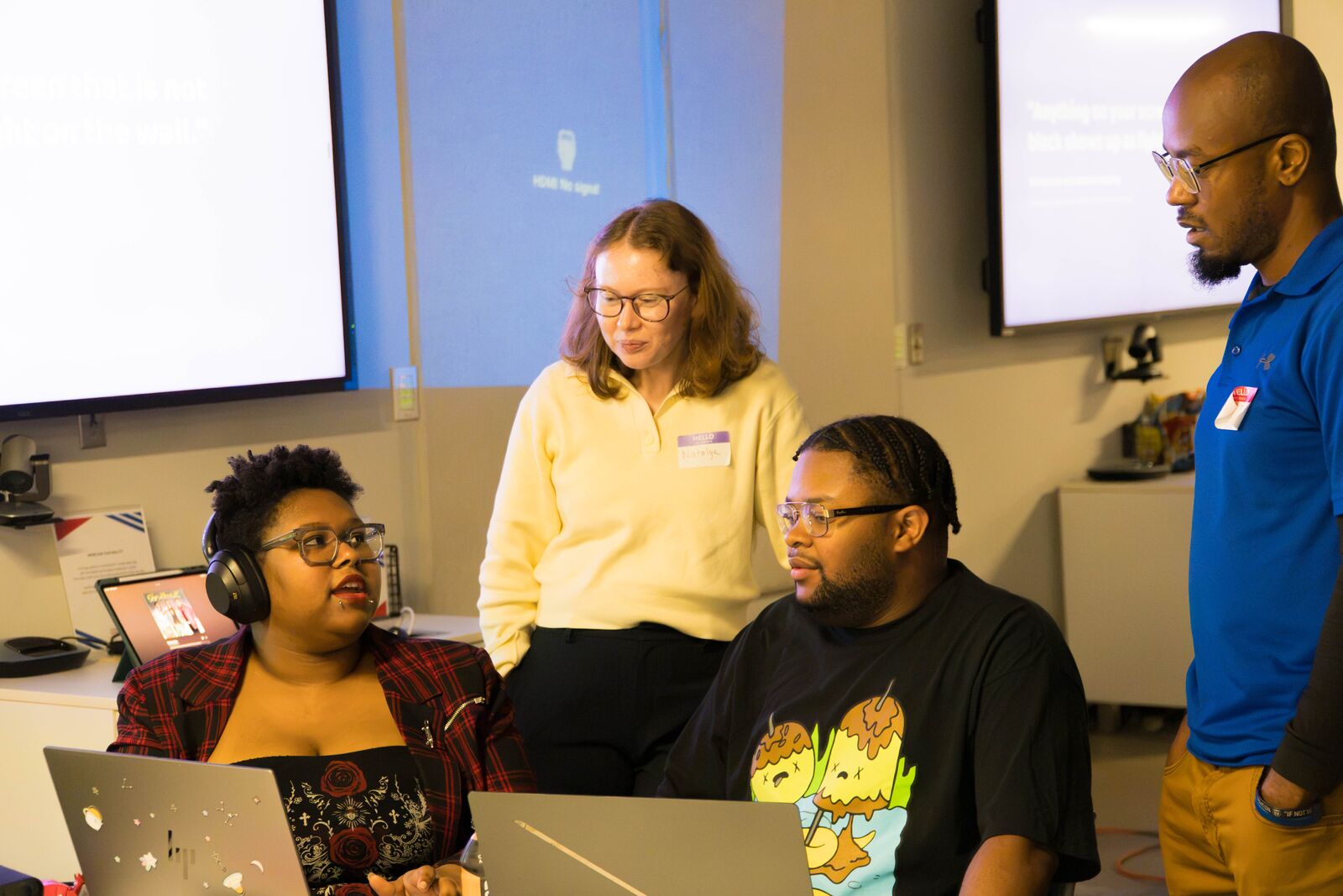Three attendees at their laptops talk with a helper standing beside them, with projection screens behind them.