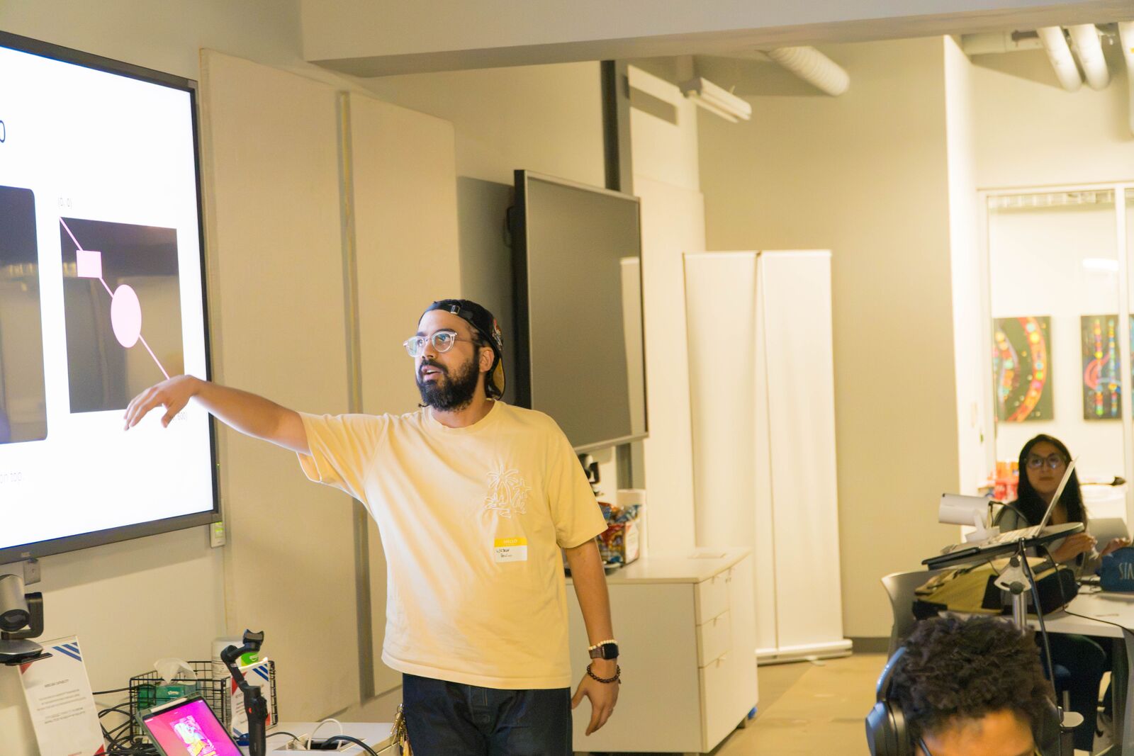 A facilitator in a cream T-shirt points at a projected slide that shows a shape with draggable corner handles, while an attendee watches from a table.